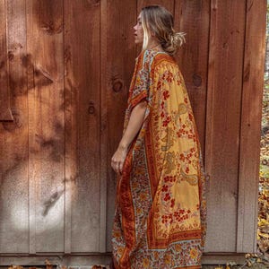 May include: A long, patterned kimono in yellow, orange, and blue. The kimono has a floral and paisley design. The garment is worn over a dark skirt. The background is a wooden wall and fallen leaves.