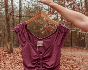 May include: A burgundy crop top with a V-neck and short sleeves. The top is made of a soft, stretchy fabric and has a gathered detail at the front. The top is hanging on a wooden hanger.