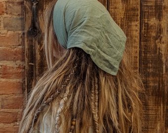 May include: A person with long, blonde dreadlocks is wearing a light green bandana. The bandana is tied around their head and covers their forehead. The person is wearing a white shirt and is standing in front of a wooden door.