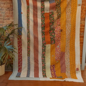 K&ouml;nnte beinhalten: Eine Patchwork-Decke mit einem gestreiften Muster in Blaut&ouml;nen, Orange, Gr&uuml;n und Wei&szlig;. Die Decke besteht aus verschiedenen Stoffen mit Blumenmustern.