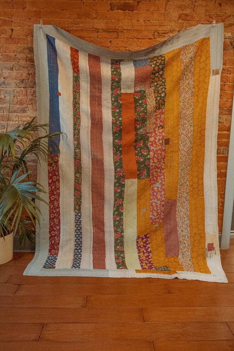 K&ouml;nnte beinhalten: Eine Patchwork-Decke mit einem gestreiften Muster in Blaut&ouml;nen, Orange, Gr&uuml;n und Wei&szlig;. Die Decke besteht aus verschiedenen Stoffen mit Blumenmustern.
