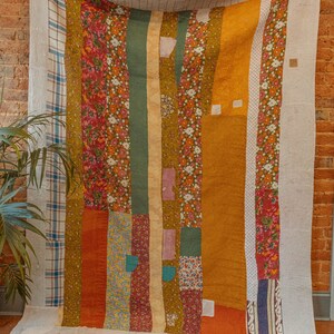May include: A patchwork quilt with a variety of floral and geometric patterns in shades of orange, brown, green, and white. The quilt is hanging on a brick wall.