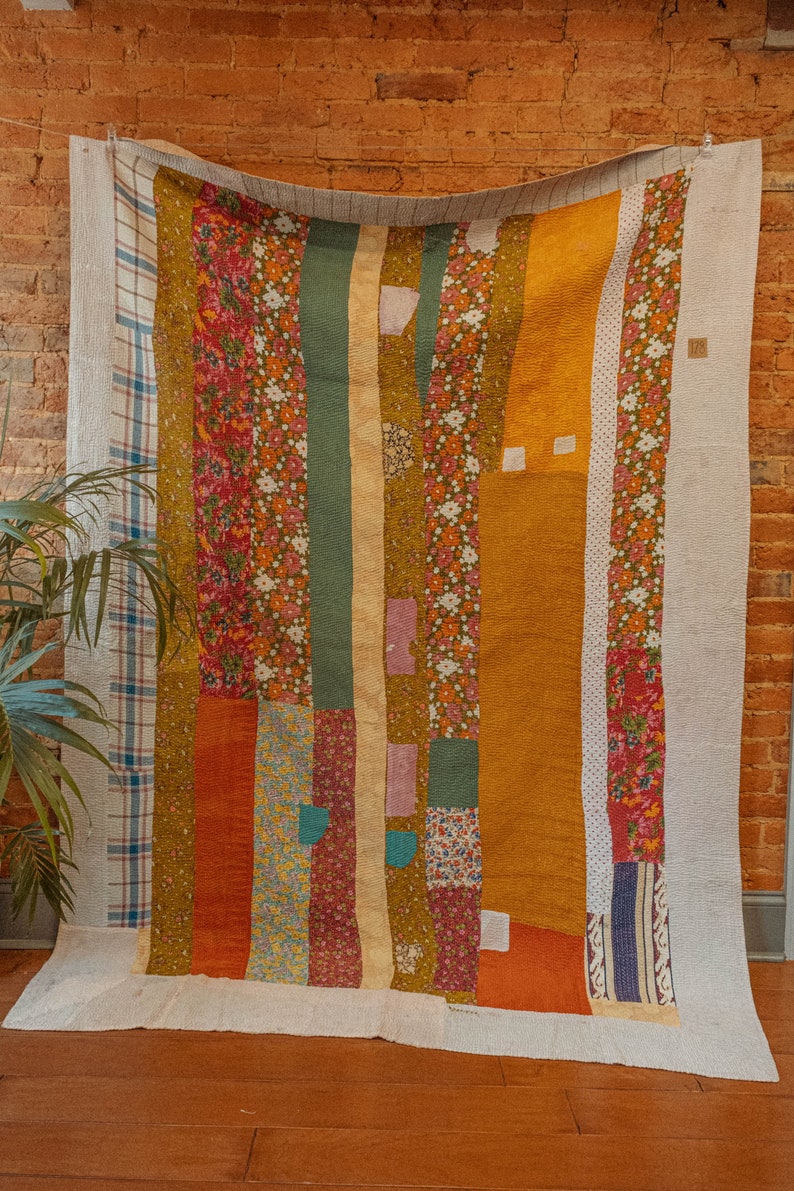 May include: A patchwork quilt with a variety of floral and geometric patterns in shades of orange, brown, green, and white. The quilt is hanging on a brick wall.