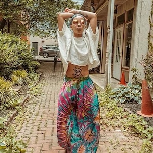 Op de afbeelding: Een persoon draagt een witte top en een kleurrijke tie-dye broek. De broek heeft een levendige mix van roze, blauwe en groene tinten. De outfit is gecombineerd met een hoofdband en zonnebril. De persoon staat op een bakstenen voetpad.
