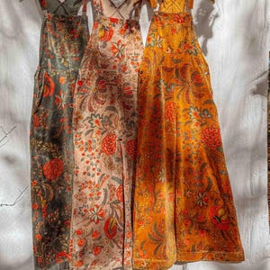 Op de afbeelding: Drie overalls met bloemenprint in olijfgroen, blush roze en goudoranje hangen aan houten hangers. De overalls hebben een wijd model, voorzakken en verstelbare bandjes. Het bloemenpatroon bevat rode, oranje en blauwe accenten.