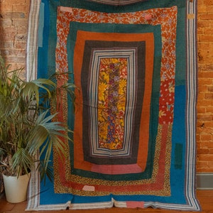 May include: A colorful patchwork quilt with a geometric design. The quilt is made of different fabrics in shades of blue, green, orange, and pink. The quilt is hanging on a brick wall.