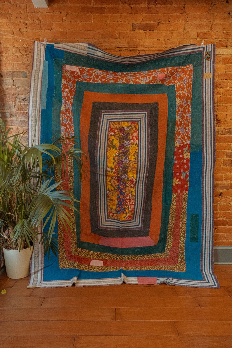 May include: A colorful patchwork quilt with a geometric design. The quilt is made of different fabrics in shades of blue, green, orange, and pink. The quilt is hanging on a brick wall.