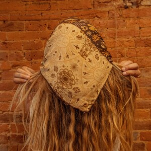 May include: A person with long, wavy, light brown hair is wearing a brown and beige bandana with a sun and floral pattern. The bandana is tied around their head and covers their forehead.