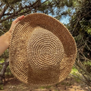 Pu&ograve; includere: Un cappello di paglia intrecciata di colore naturale, con tesa larga, su uno sfondo di fogliame verde e cielo blu. Il cappello presenta un motivo circolare sulla corona e una tesa leggermente curva. Un accessorio estivo.