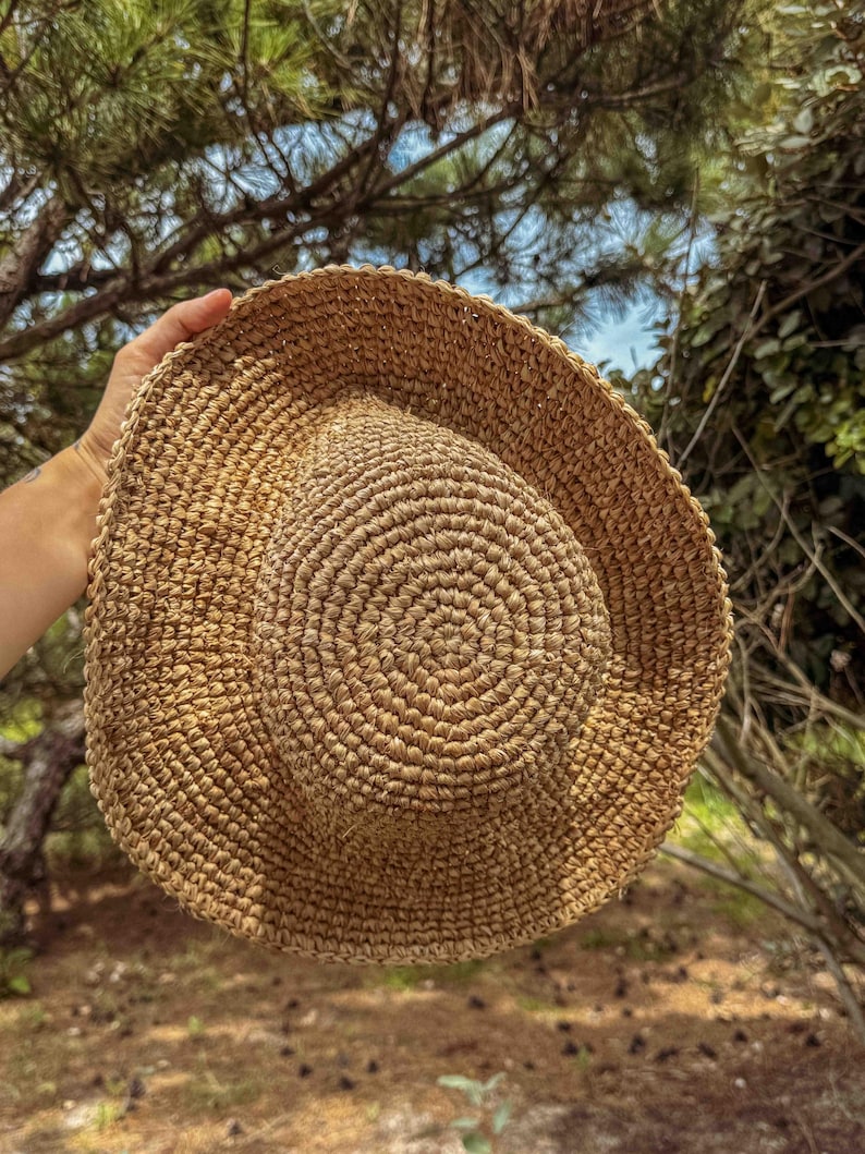 Pu&ograve; includere: Un cappello di paglia intrecciata di colore naturale, con tesa larga, su uno sfondo di fogliame verde e cielo blu. Il cappello presenta un motivo circolare sulla corona e una tesa leggermente curva. Un accessorio estivo.