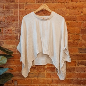 May include: A white, lightweight, cotton top with a relaxed fit and a slightly cropped hem. The top is hanging on a wooden hanger against a brick wall.