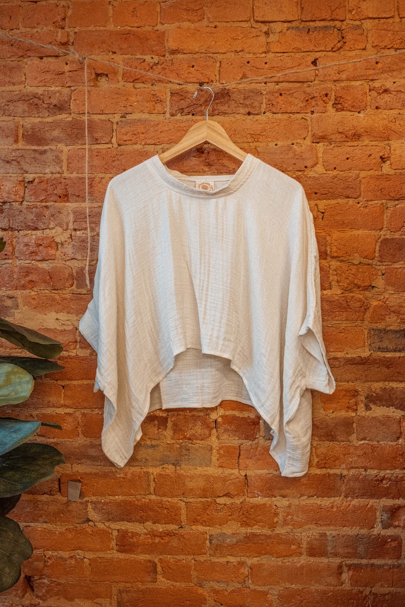 May include: A white, lightweight, cotton top with a relaxed fit and a slightly cropped hem. The top is hanging on a wooden hanger against a brick wall.