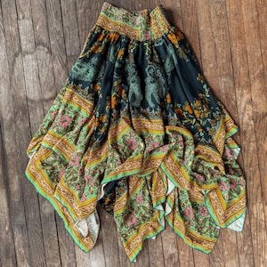 May include: A flowing, patterned skirt with a dark background and a vibrant floral and animal print. The skirt features a mix of green, orange, pink, and gold colors, with a wide, elasticated waistband. The fabric drapes elegantly, showcasing the intricate design.