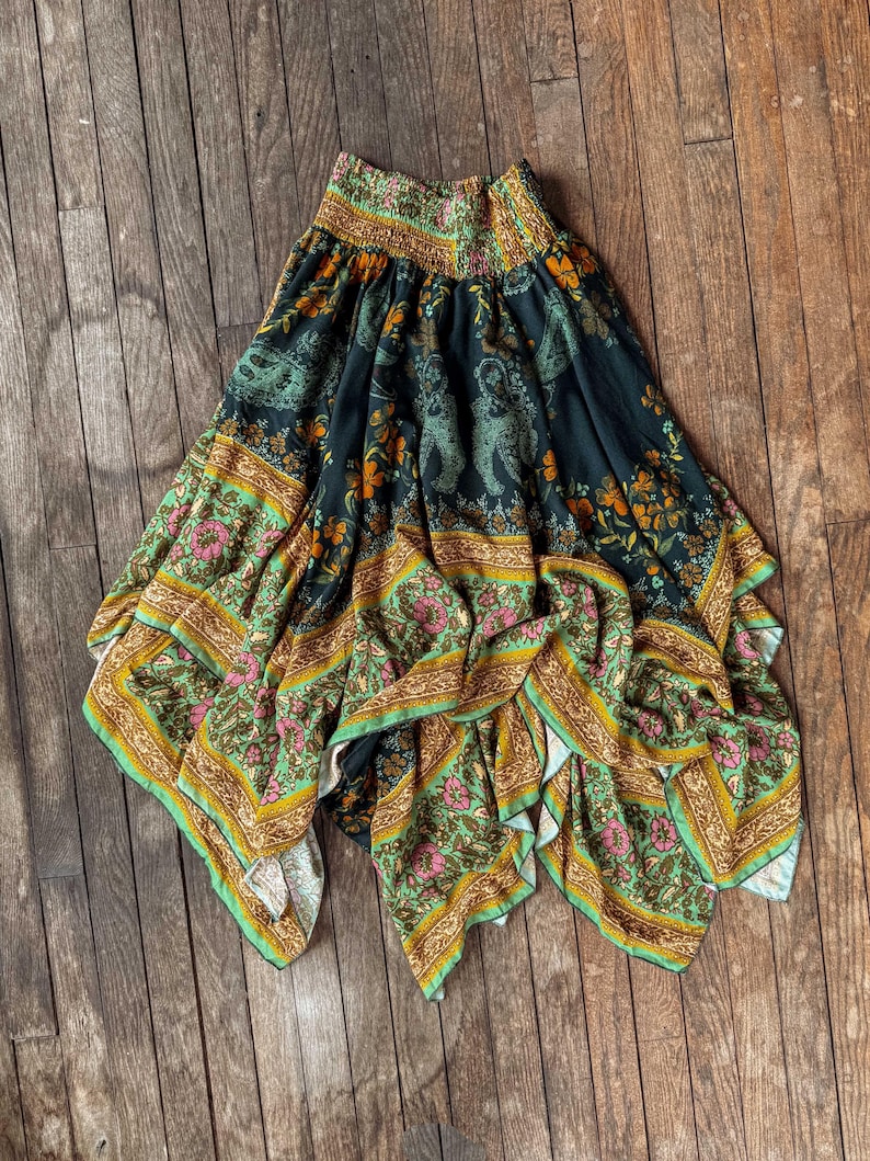 May include: A flowing, patterned skirt with a dark background and a vibrant floral and animal print. The skirt features a mix of green, orange, pink, and gold colors, with a wide, elasticated waistband. The fabric drapes elegantly, showcasing the intricate design.
