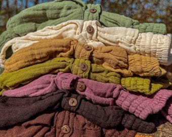 May include: A stack of button-down shirts in various colors, including green, cream, mustard, and brown. Each shirt has a textured fabric and wooden buttons. The shirts are neatly arranged, showcasing a range of earthy tones.