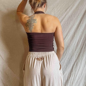 K&ouml;nnte beinhalten: Ein dunkelbraunes Neckholder-Top kombiniert mit einer hellbeigen Hose mit weitem Bein. Die Hose hat einen elastischen Bund und ein kleines, gesticktes Motten-Design in Braun- und Goldt&ouml;nen, das mittig unter dem Bund auf der R&uuml;ckseite platziert ist.