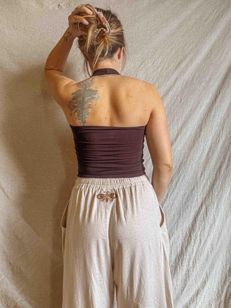 K&ouml;nnte beinhalten: Ein dunkelbraunes Neckholder-Top kombiniert mit einer hellbeigen Hose mit weitem Bein. Die Hose hat einen elastischen Bund und ein kleines, gesticktes Motten-Design in Braun- und Goldt&ouml;nen, das mittig unter dem Bund auf der R&uuml;ckseite platziert ist.