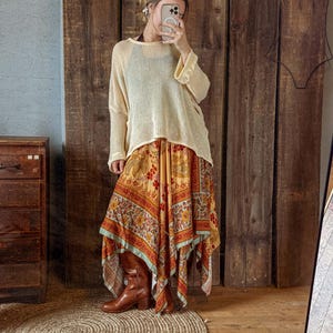 May include: A woman's outfit featuring a cream-colored, long-sleeved knit top and a flowing, patterned skirt in shades of yellow, orange, and red. The skirt has a unique asymmetrical hemline. Brown leather boots complete the look.