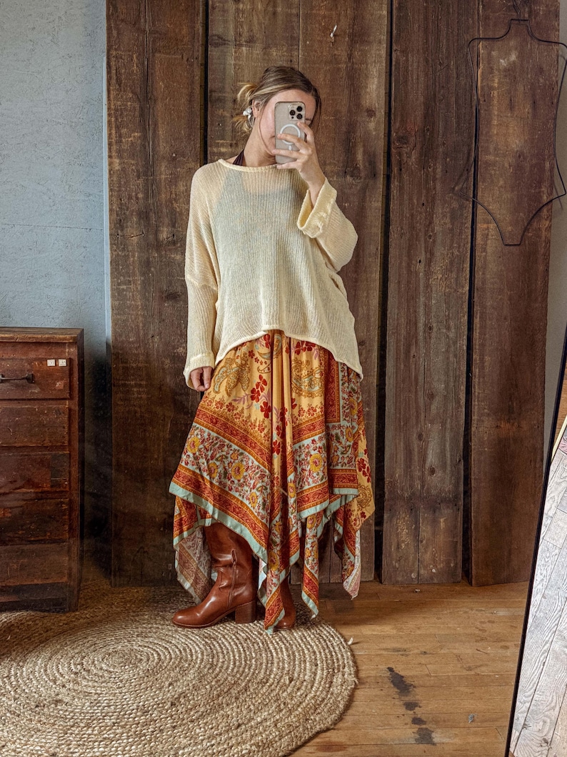 May include: A woman's outfit featuring a cream-colored, long-sleeved knit top and a flowing, patterned skirt in shades of yellow, orange, and red. The skirt has a unique asymmetrical hemline. Brown leather boots complete the look.