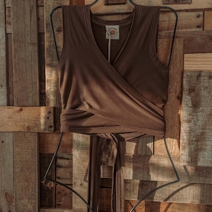 May include: A brown sleeveless wrap top with a V-neckline. The top is tied at the waist and has a small brown tag with the text "SUNDANCE" on it.