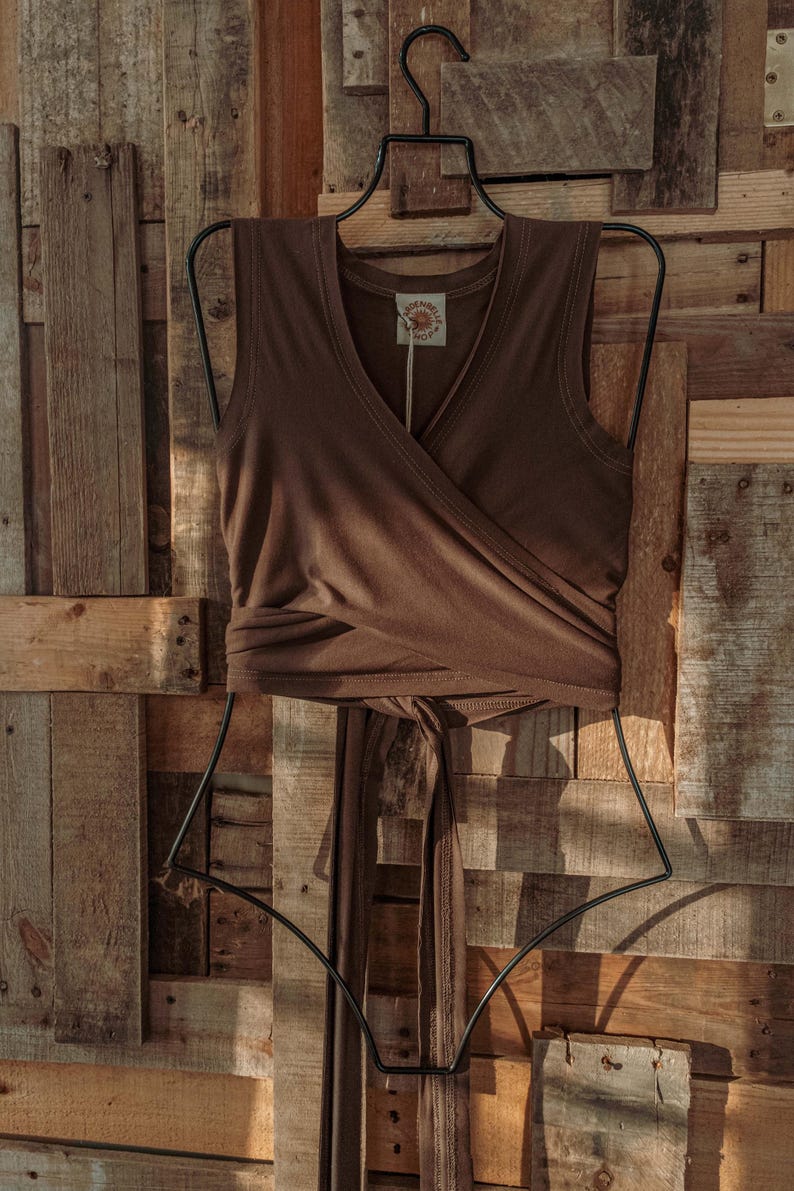 May include: A brown sleeveless wrap top with a V-neckline. The top is tied at the waist and has a small brown tag with the text "SUNDANCE" on it.