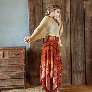 May include: A woman's outfit featuring a cream-colored, long-sleeved sweater and a long, flowing skirt. The skirt has a vibrant, colorful pattern with shades of red, orange, and pink. The outfit is styled in a bohemian fashion.
