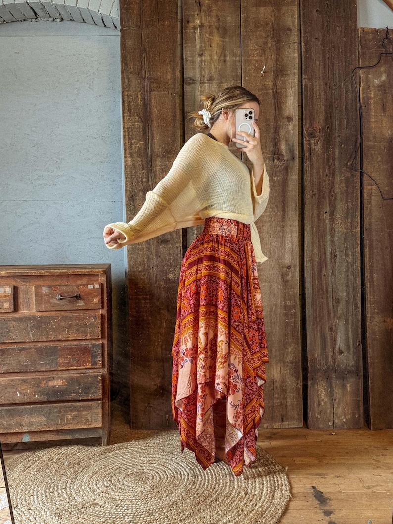 May include: A woman's outfit featuring a cream-colored, long-sleeved sweater and a long, flowing skirt. The skirt has a vibrant, colorful pattern with shades of red, orange, and pink. The outfit is styled in a bohemian fashion.