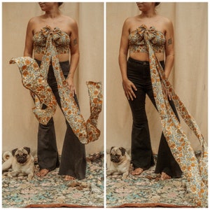 May include: Two women wearing a floral print top with a matching scarf tied around their waist. The top is a halter style and the scarf is tied in a bow at the front. The women are wearing black flared pants and are standing in front of a patterned rug. A pug dog is sitting in front of each woman.