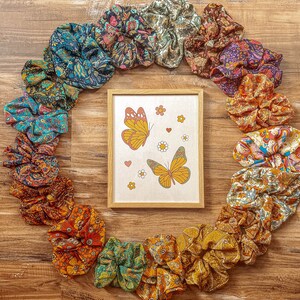 May include: A circle of colorful patterned hair scrunchies with a framed print of two butterflies in the center. The butterflies are orange and yellow with black markings and are flying against a white background with pink and white flowers.