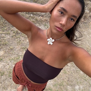 May include: A woman wearing a dark brown tube top and rust-colored pants. A flower-shaped shell necklace is around her neck. The background is a field of dry grass.