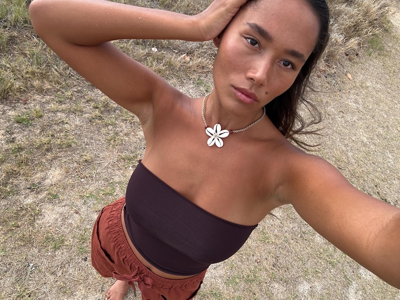 May include: A woman wearing a dark brown tube top and rust-colored pants. A flower-shaped shell necklace is around her neck. The background is a field of dry grass.