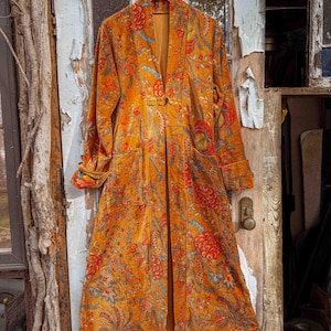 May include: An orange, long-sleeved coat with a floral pattern in red, blue, and yellow. The coat has a single button closure and two front pockets. It is hanging on a wooden hanger.