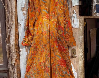 Puede incluir: Un abrigo largo naranja con un estampado floral en rojo, azul y amarillo. El abrigo tiene mangas largas, un solo botón y dos bolsillos. Cuelga de una percha de madera.