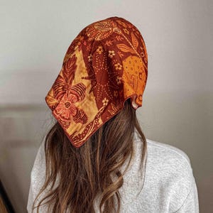Puede incluir: Un pa&ntilde;uelo estampado en tonos naranja, marr&oacute;n y dorado. El pa&ntilde;uelo presenta un dise&ntilde;o floral y se lleva en la cabeza. El pa&ntilde;uelo est&aacute; hecho de un material suave y es un accesorio de moda.