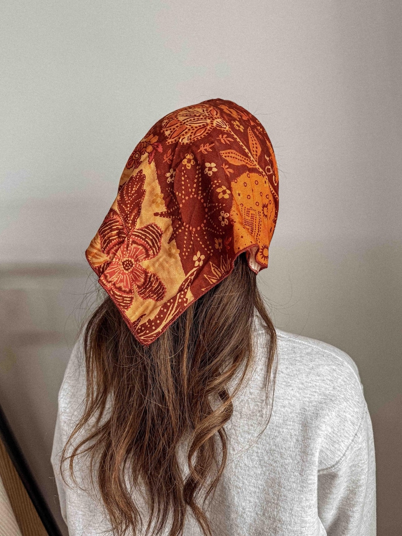 Puede incluir: Un pa&ntilde;uelo estampado en tonos naranja, marr&oacute;n y dorado. El pa&ntilde;uelo presenta un dise&ntilde;o floral y se lleva en la cabeza. El pa&ntilde;uelo est&aacute; hecho de un material suave y es un accesorio de moda.