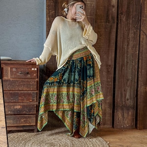 May include: A cream-colored, oversized sweater paired with a long, flowing skirt. The skirt features a dark teal background with a colorful floral and paisley pattern. The skirt has a unique asymmetrical hemline.