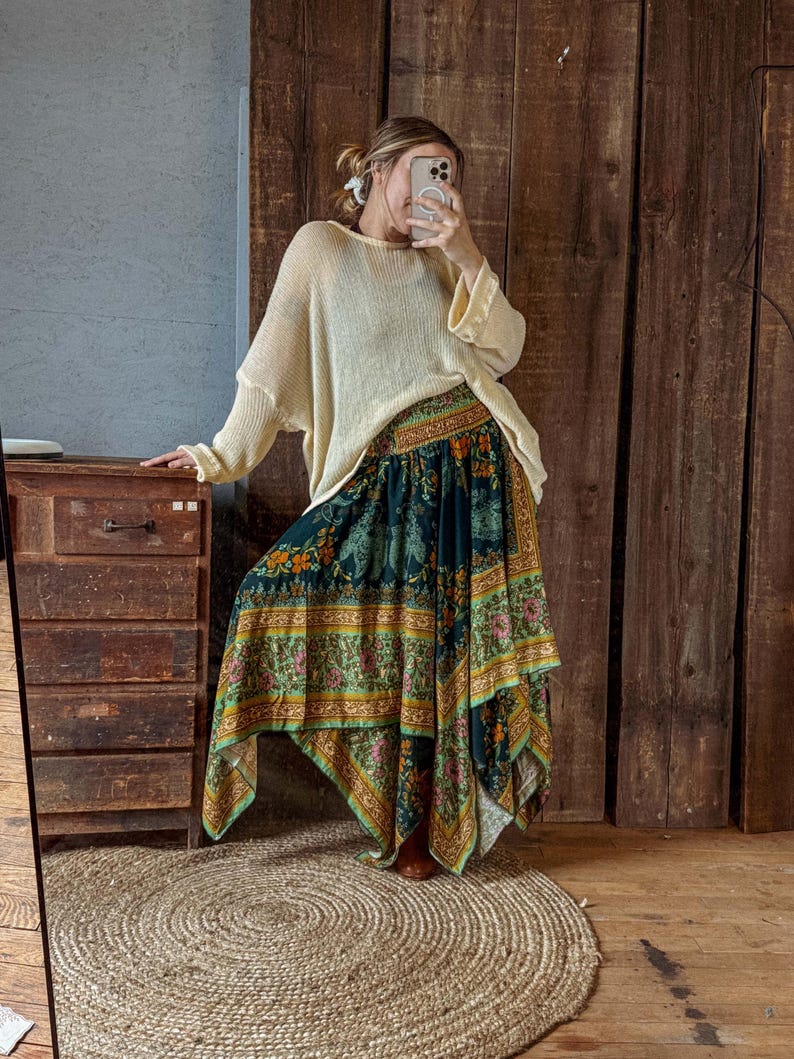 May include: A cream-colored, oversized sweater paired with a long, flowing skirt. The skirt features a dark teal background with a colorful floral and paisley pattern. The skirt has a unique asymmetrical hemline.