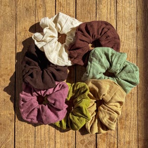 May include: A set of eight fabric scrunchies in various colors, including brown, beige, green, and purple. The scrunchies are arranged in a circle on a wooden surface.