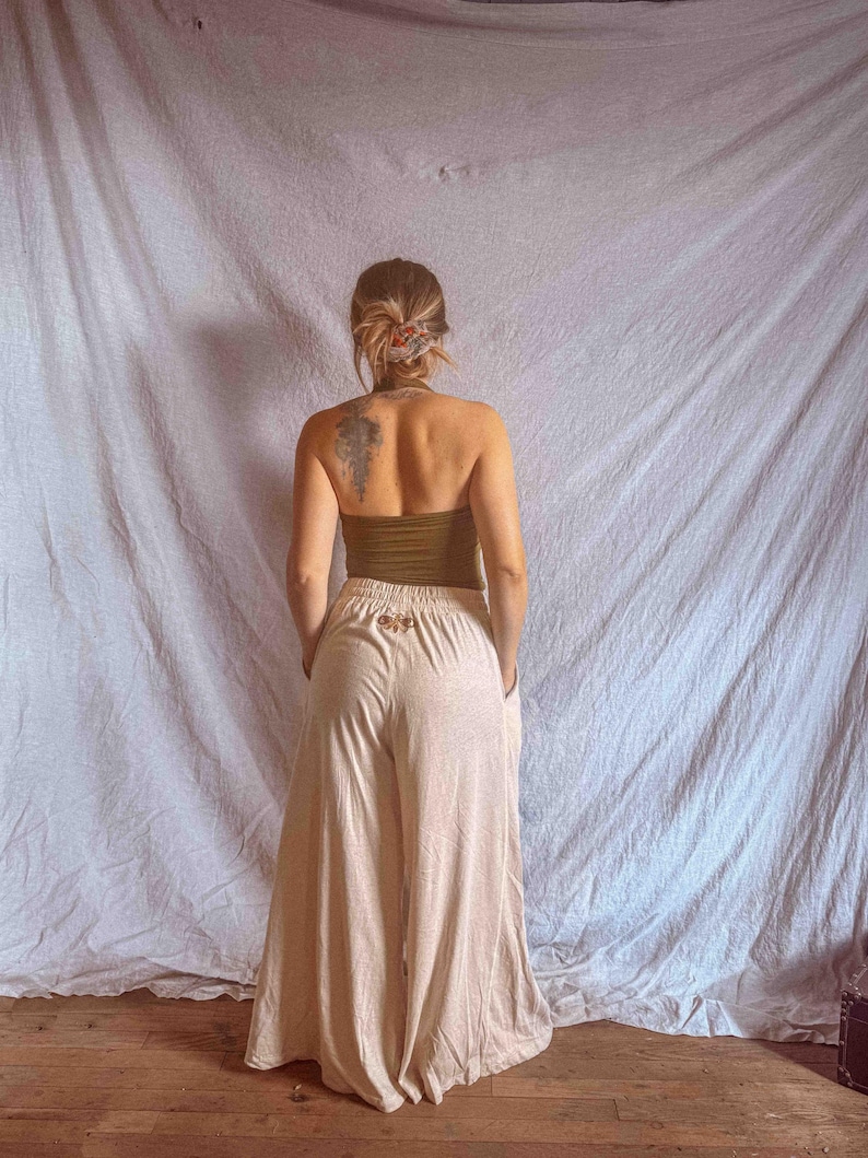 K&ouml;nnte beinhalten: Eine hoch taillierte Hose mit weitem Bein in hellem Beige, die ein kleines gesticktes Mottenmotiv am hinteren Bund aufweist. Die Hose hat eine lockere, flie&szlig;ende Silhouette mit Seitentaschen und einem elastischen Bund.