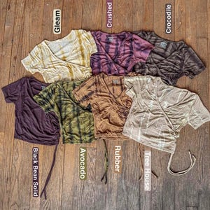 Puede incluir: Una colección de tops cruzados de manga corta en varios colores, incluyendo amarillo, morado, marrón, verde y beige. Cada top tiene un cuello en V y está etiquetado con un nombre de color. Los tops están colocados sobre una superficie de madera.