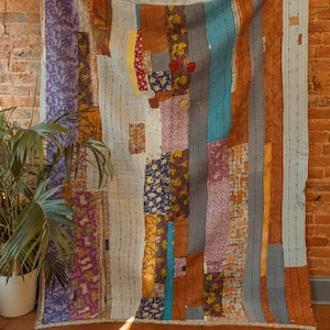 Op de afbeelding: Een patchworkdeken met een verscheidenheid aan kleuren en patronen, waaronder blauw, paars, bruin en geel. De deken is gemaakt van verschillende stoffen die in een willekeurig patroon aan elkaar zijn genaaid. Het hangt aan een bakstenen muur.