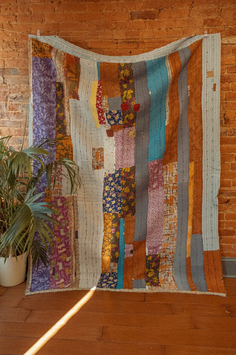 Op de afbeelding: Een patchworkdeken met een verscheidenheid aan kleuren en patronen, waaronder blauw, paars, bruin en geel. De deken is gemaakt van verschillende stoffen die in een willekeurig patroon aan elkaar zijn genaaid. Het hangt aan een bakstenen muur.