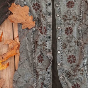 May include: A gray denim vest with a black fabric lining and floral embroidery. The vest has a button closure and a ruffled trim.