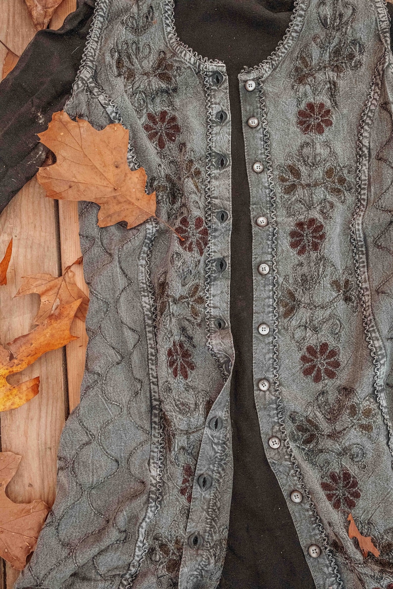 May include: A gray denim vest with a black fabric lining and floral embroidery. The vest has a button closure and a ruffled trim.