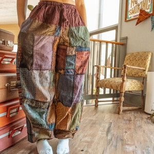 May include: A person wearing a white crop top and patchwork pants. The pants are made of different colored fabrics and have a loose fit. The person is standing on a wooden floor.