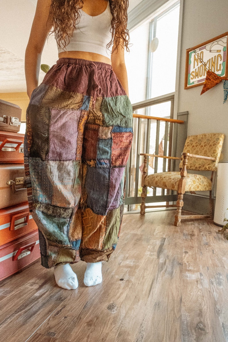 May include: A person wearing a white crop top and patchwork pants. The pants are made of different colored fabrics and have a loose fit. The person is standing on a wooden floor.