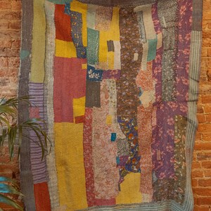K&ouml;nnte beinhalten: Eine bunte Patchwork-Decke mit verschiedenen floralen und geometrischen Mustern in Rot-, Gelb-, Lila- und Gr&uuml;nt&ouml;nen. Die Decke h&auml;ngt an einer Backsteinwand.