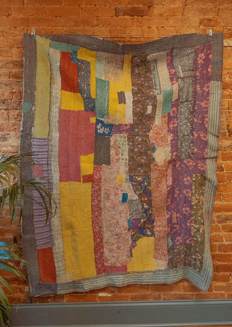 K&ouml;nnte beinhalten: Eine bunte Patchwork-Decke mit verschiedenen floralen und geometrischen Mustern in Rot-, Gelb-, Lila- und Gr&uuml;nt&ouml;nen. Die Decke h&auml;ngt an einer Backsteinwand.