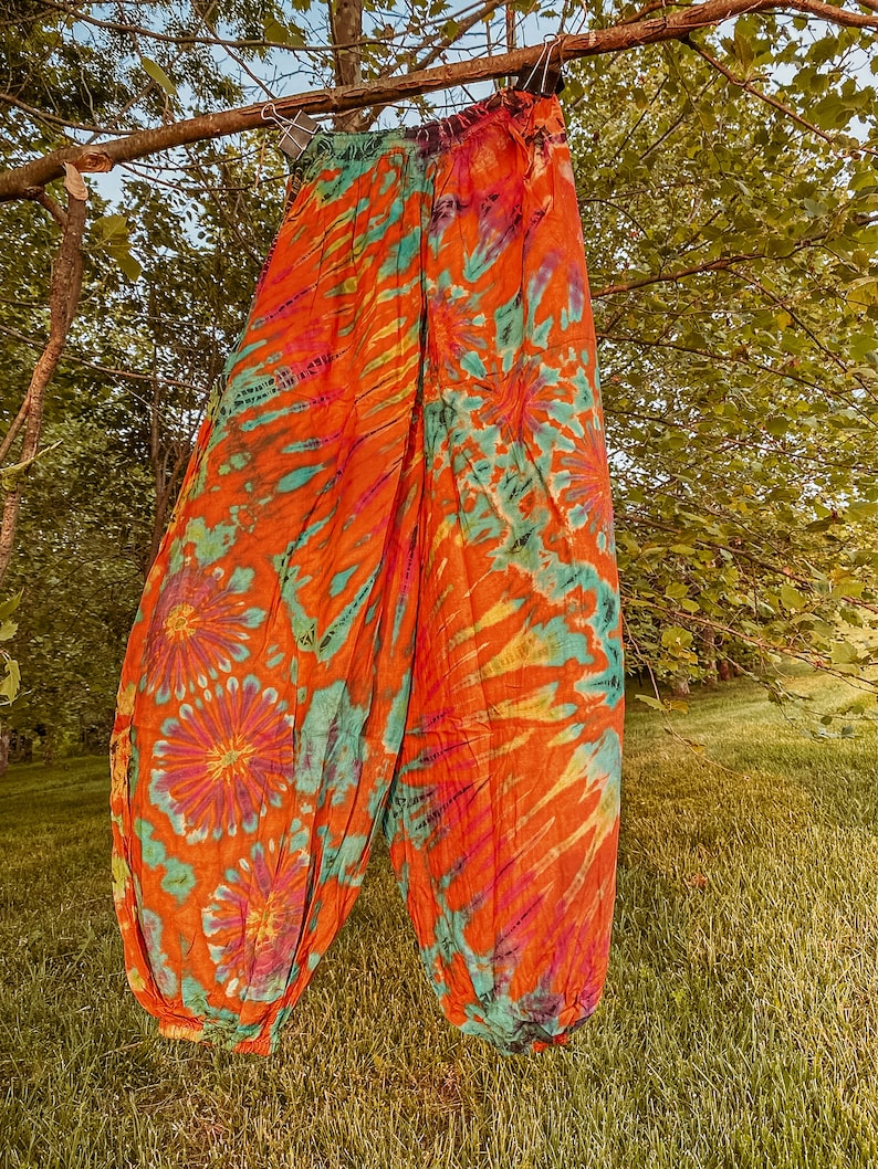 May include: A pair of tie-dye trousers in orange, teal, and purple hangs from a tree branch. The trousers have a floral pattern and a loose fit, suitable for casual wear. The fabric looks lightweight and flowing, ideal for warmer weather.