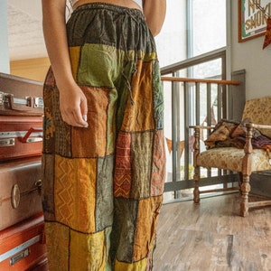 May include: A person wearing a white crop top and patchwork pants with a drawstring waist. The pants are made of different fabrics and colors, including brown, green, yellow, and orange. The person is standing in a room with wooden floors and a chair in the background.